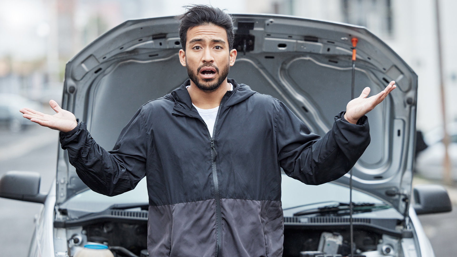 man standing in front of his vehicle with the hood open; his arms are raised in a shrug and his expression is disbelief.
